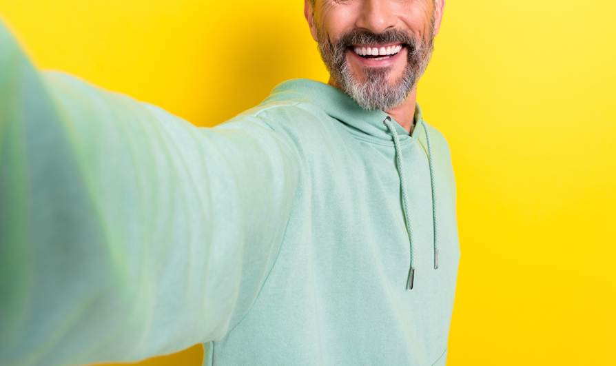 Halve ansiktet til en smilende mann med grått skjegg som holder armen ut og tar en «selfie» foran gul bakgrunn.