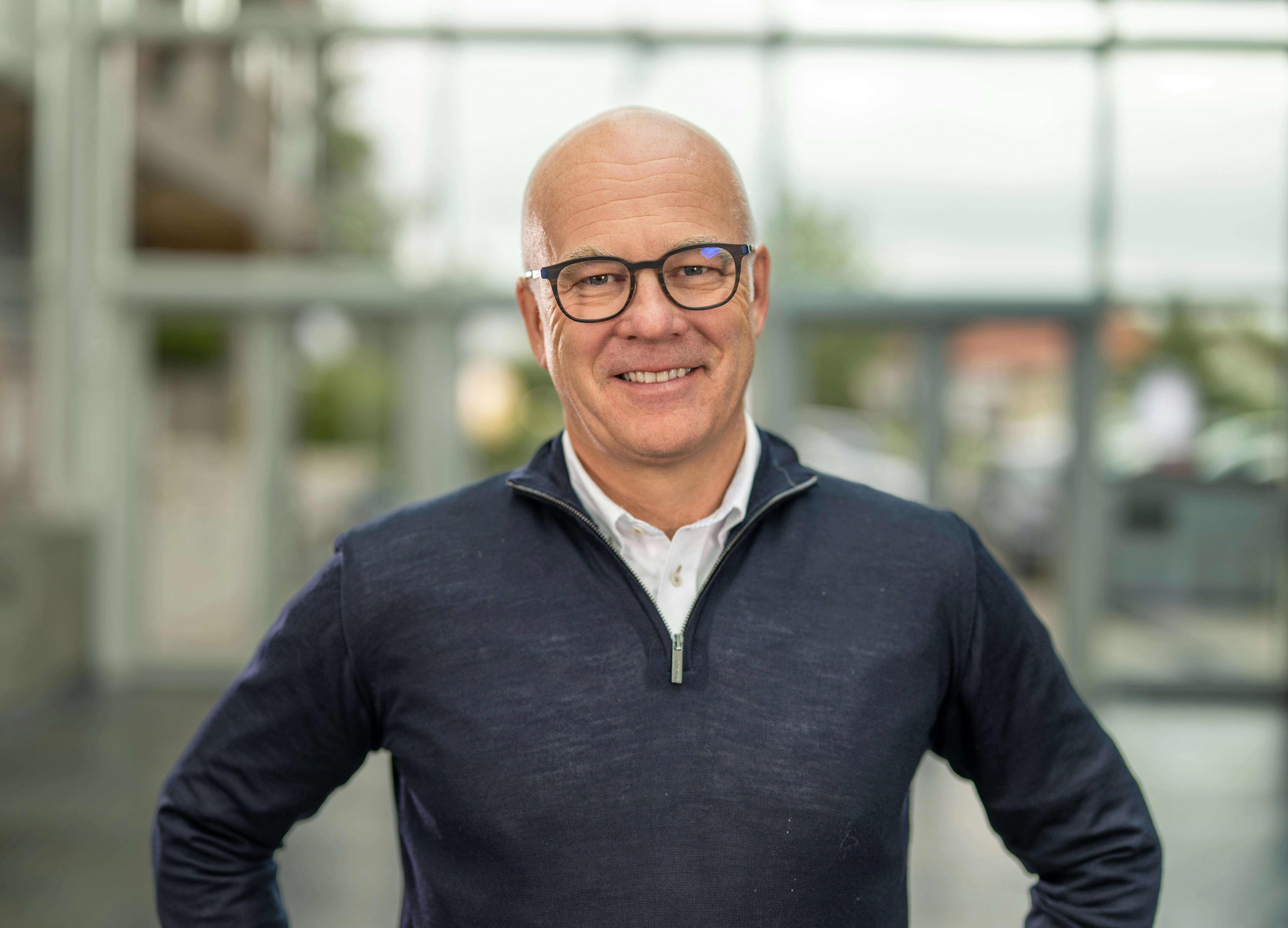 En smilende, skallet mann med briller, iført blå genser med hvit skjorte under, avbildet foran en glassvegg.