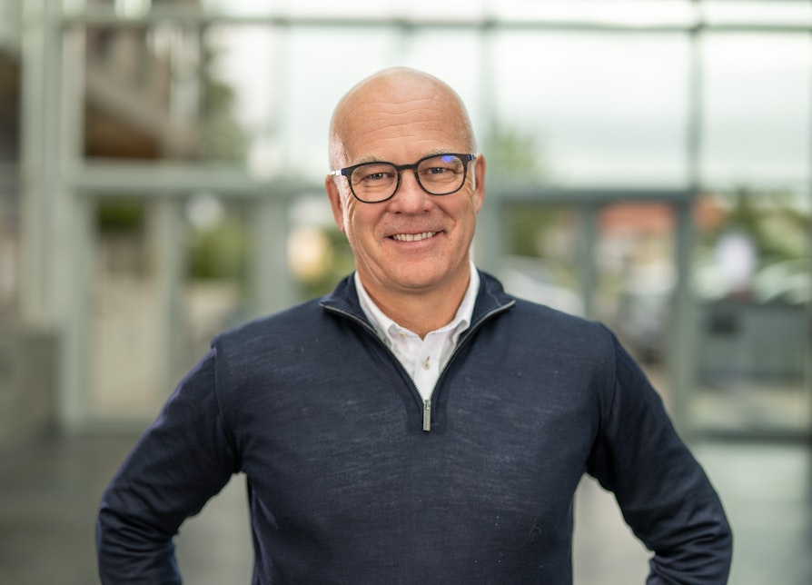 En smilende, skallet mann med briller, iført blå genser med hvit skjorte under, avbildet foran en glassvegg.