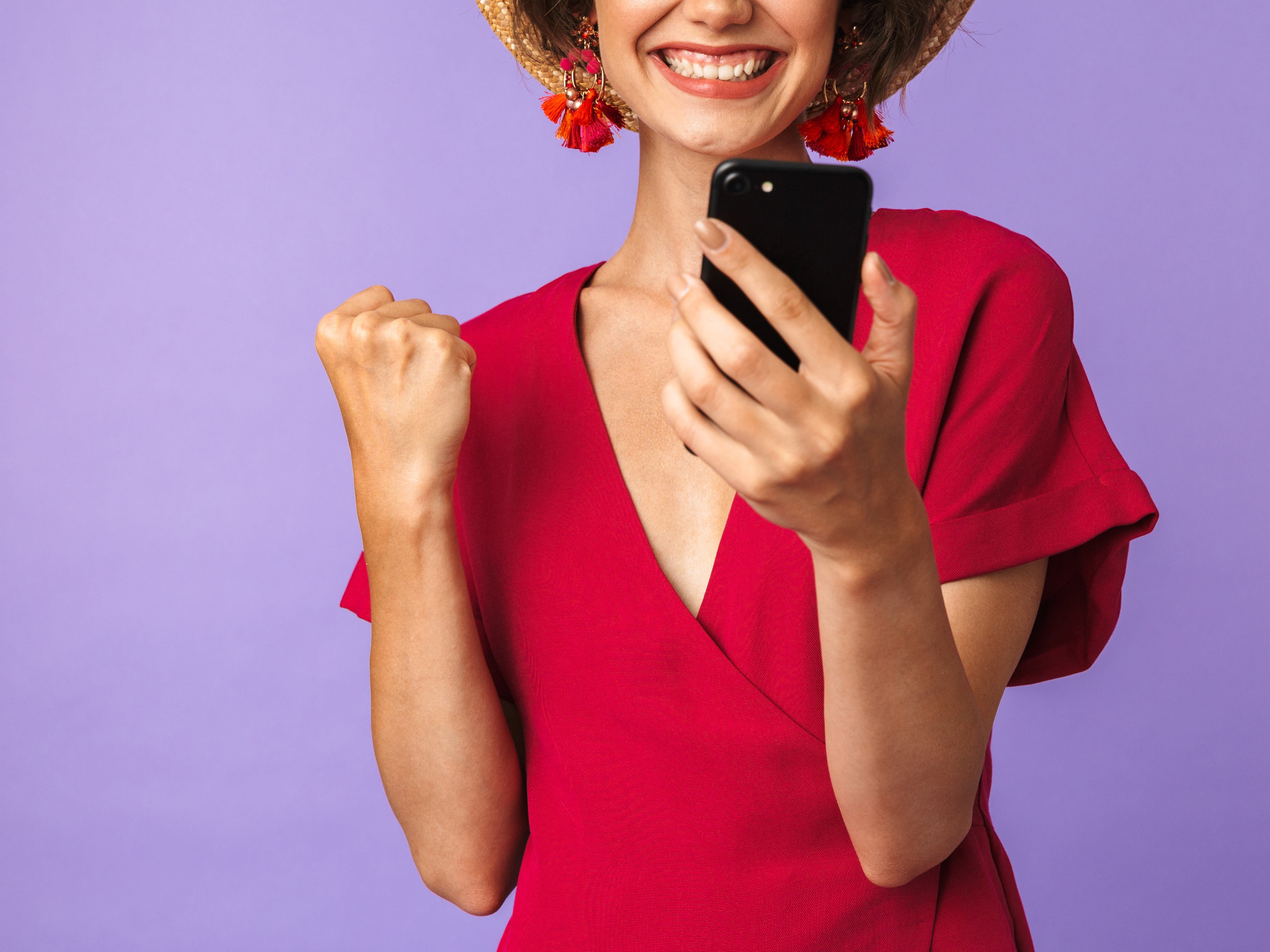 Kvinne i rød kjole og hatt ser på en mobil mens hun smiler og knytter den ene neven i seier.