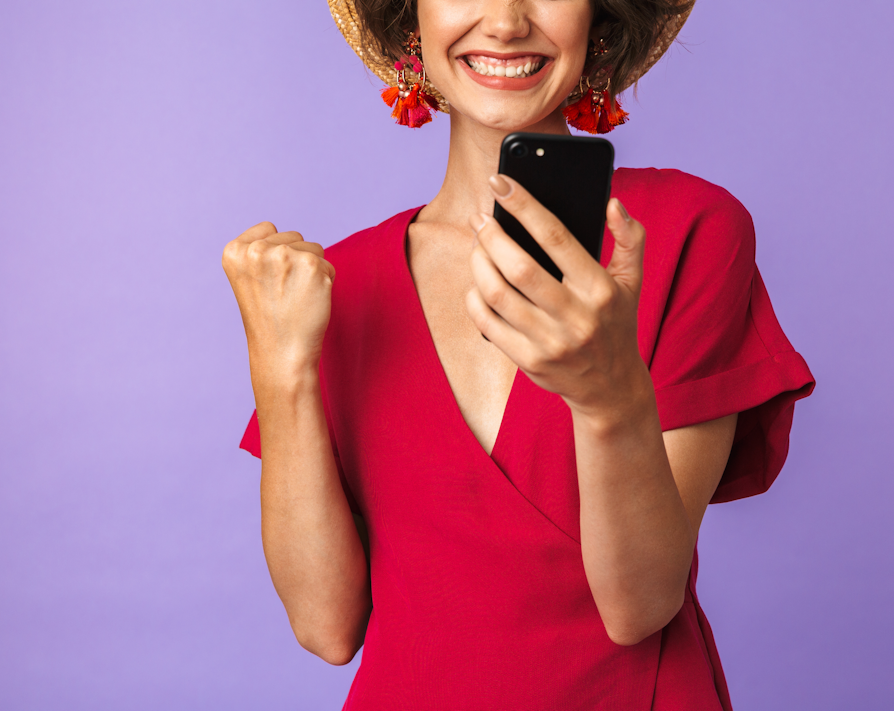 Kvinne i rød kjole og hatt ser på en mobil mens hun smiler og knytter den ene neven i seier.