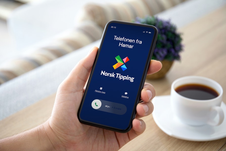 Mannehånd holder mobiltelefon som ringer fra Telefonen fra Hamar over et bord med en kaffekopp og en blomst.