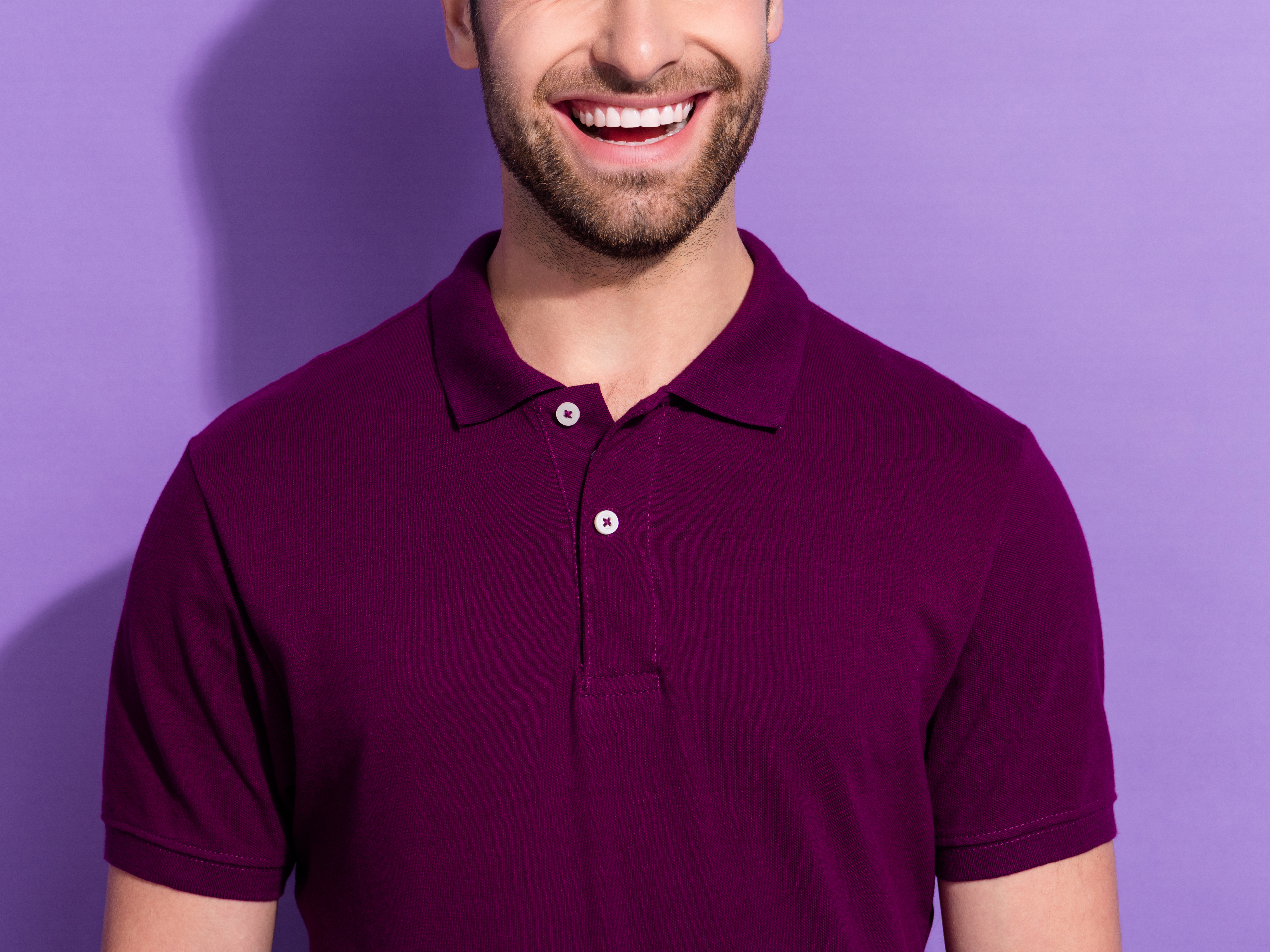 Mann som smiler med skjegg i lilla t-skjorte med krage mot lilla bakgrunn.