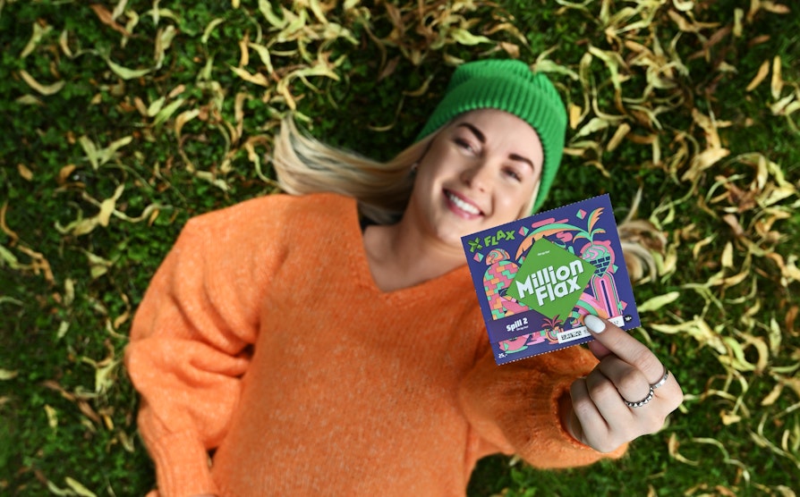 Blond kvinne med grønn lue og oransje genser ligger smilende på gresset med gult løv mens hun holder opp et MillionFlax-lodd.