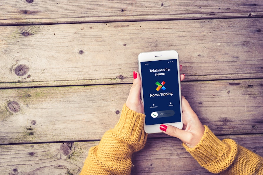 Kvinnehender med rød neglelak og oransje genser holder hvit mobiltelefon som viser anrop og Telefonen fra Hamar, mot et trebord