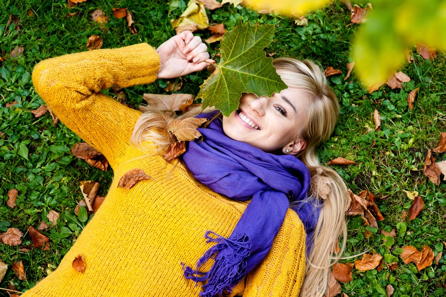 Kvinne med blondt langt hår, lilla skjerf og gul genser ligger på gress med høstløv og holder et grønt blad over det ene øyet mens hun smiler.