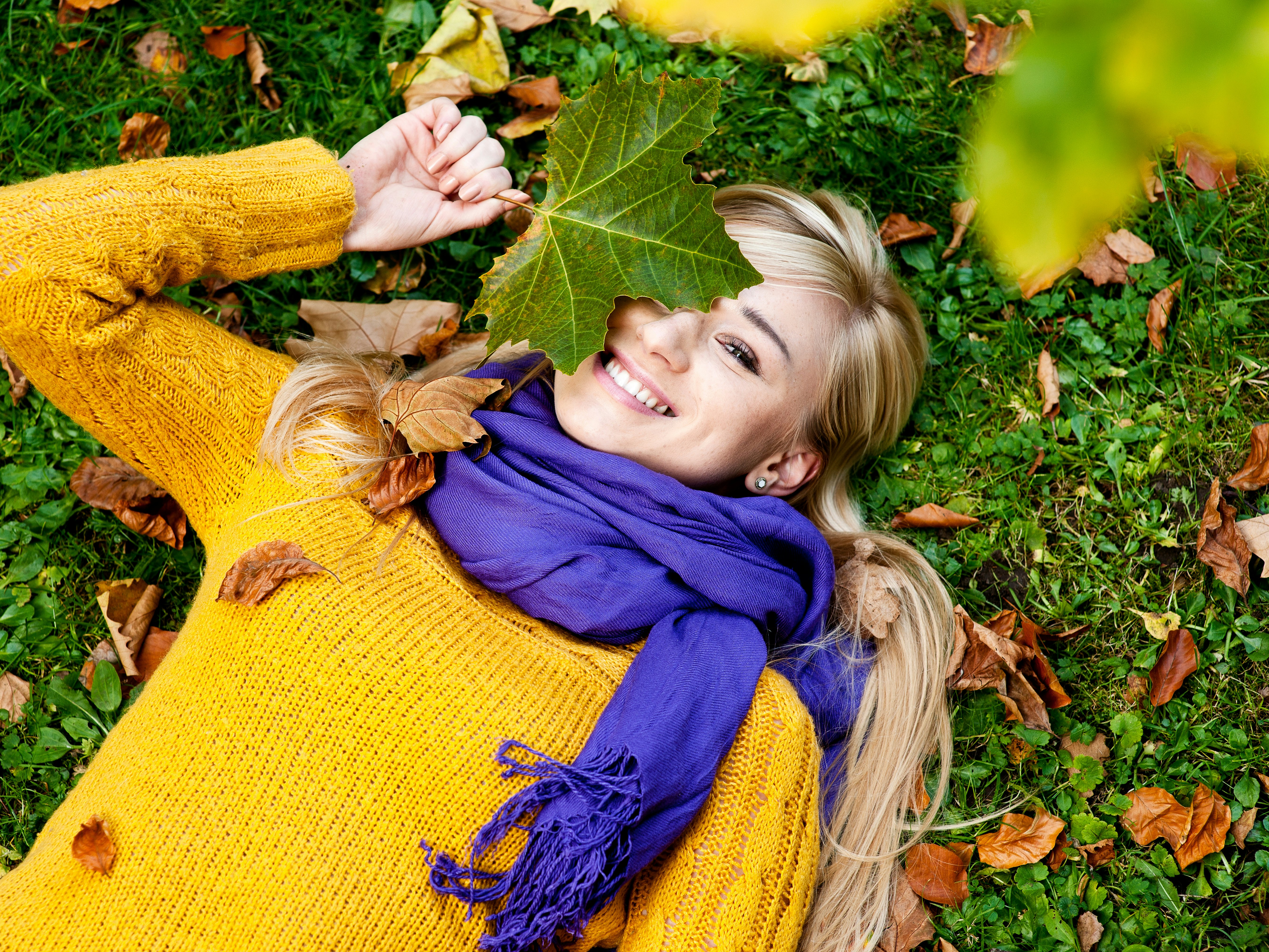 Kvinne med blondt langt hår, lilla skjerf og gul genser ligger på gress med høstløv og holder et grønt blad over det ene øyet mens hun smiler.