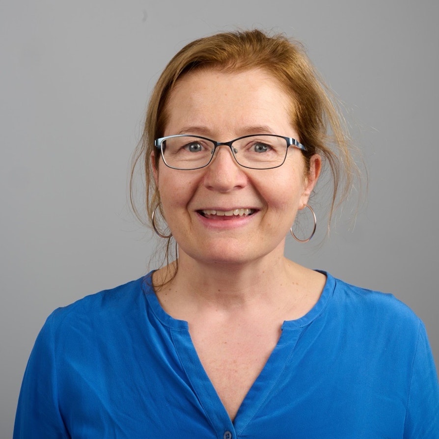 Blid kvinne på 56 år med hestehale, briller, store øreringer og kongeblå bluse.