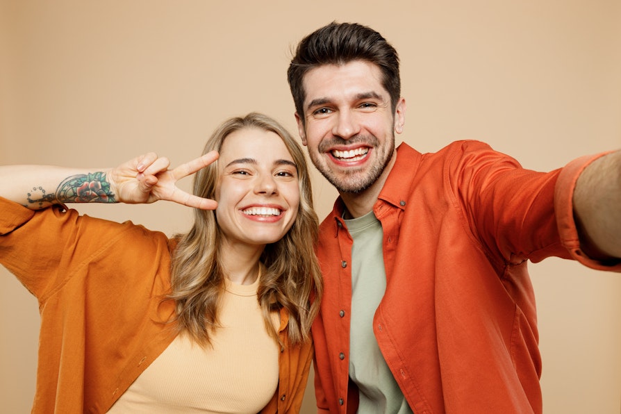 Ung blond kvinne og brunhåret mann smiler mot kamera mens de holder rundt hverandre. Begge i oransje skjorter mot ferskenfarget bakgrunn, mens kvinnen viser fredstegn.