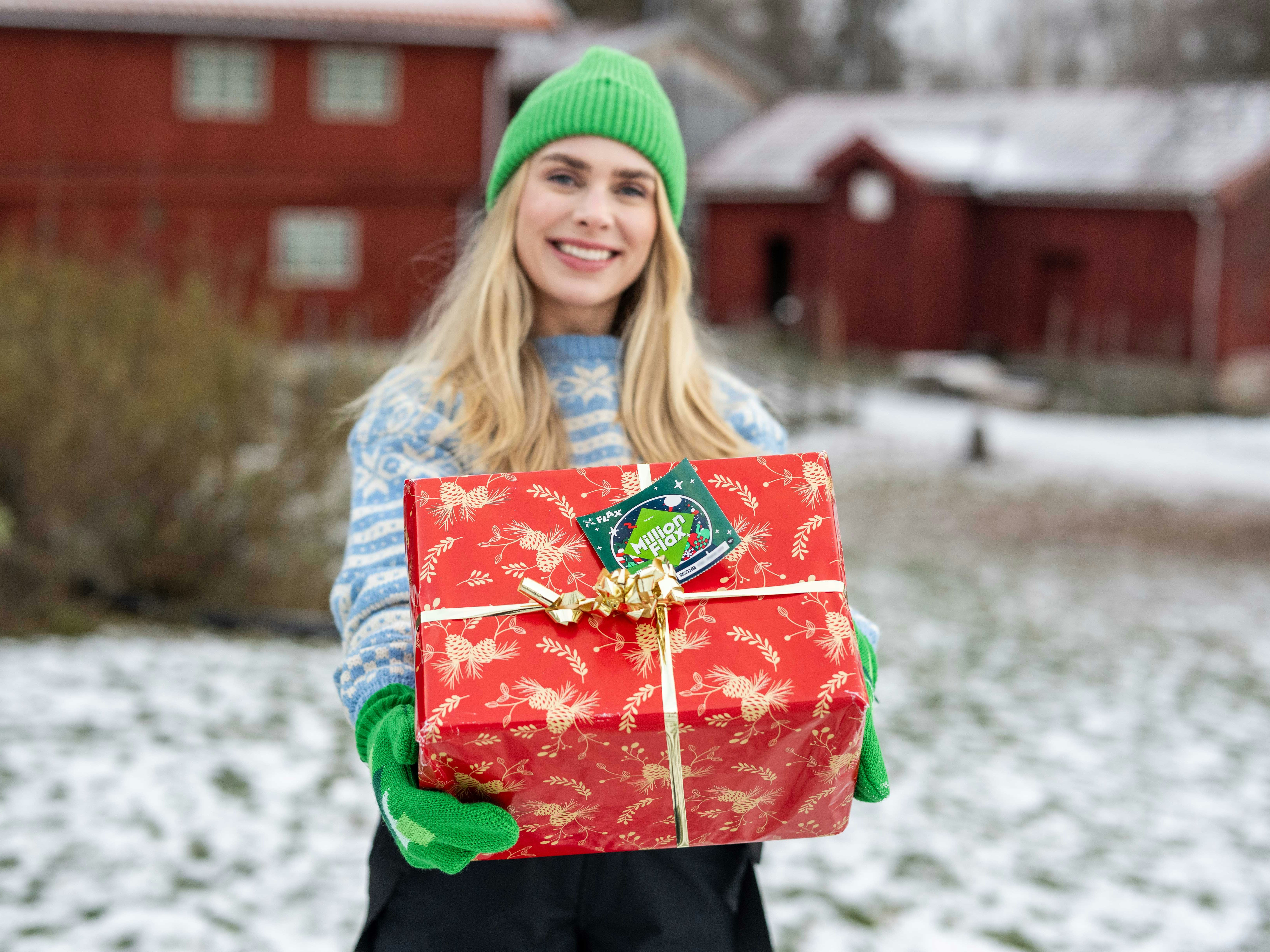 Blond  smilende kvinne med lyseblå strikkejakke og grønne votter og lue, står foran et gammelt gårdstun med snø holder frem en rød  pakke med et MillionFlax-lodd festet som gavelapp.