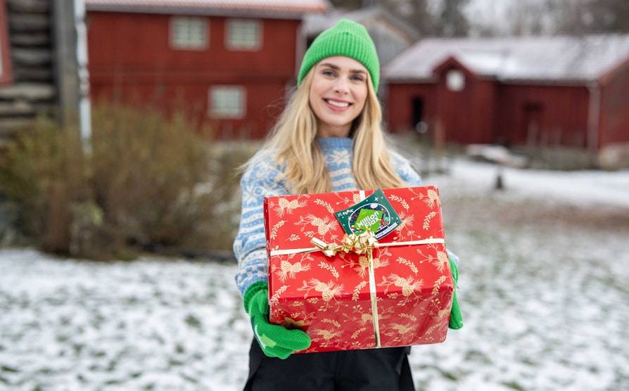 Blond  smilende kvinne med lyseblå strikkejakke og grønne votter og lue, står foran et gammelt gårdstun med snø holder frem en rød  pakke med et MillionFlax-lodd festet som gavelapp.