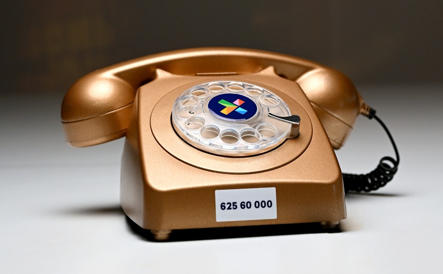 Bilde av gammeldags telefon, med dreieskive. Gullfarget. Norsk Tipping-logo + telefonnummeret 62560000.