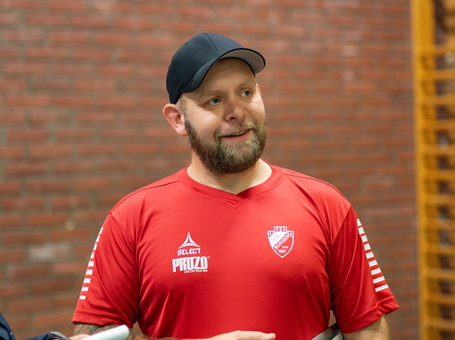 Blid mann med skjegg, svart caps og rød t-skjorte i gymsal.
