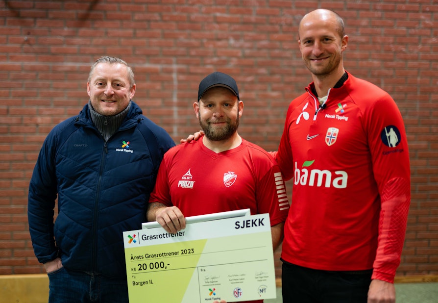 Bilde av tre glade menn. Mannen i midten har nettopp blitt overrasket og mottatt en sjekk på 20000 kroner etter å ha blitt Årets grasrottrener.