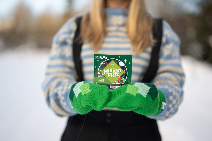 Utsnitt av en kvinneoverkropp i lyseblå mønstrete strikkejakke, sort overtrekksbukse med seler og grønne votter som holder et MillionFlax-lodd foran seg.