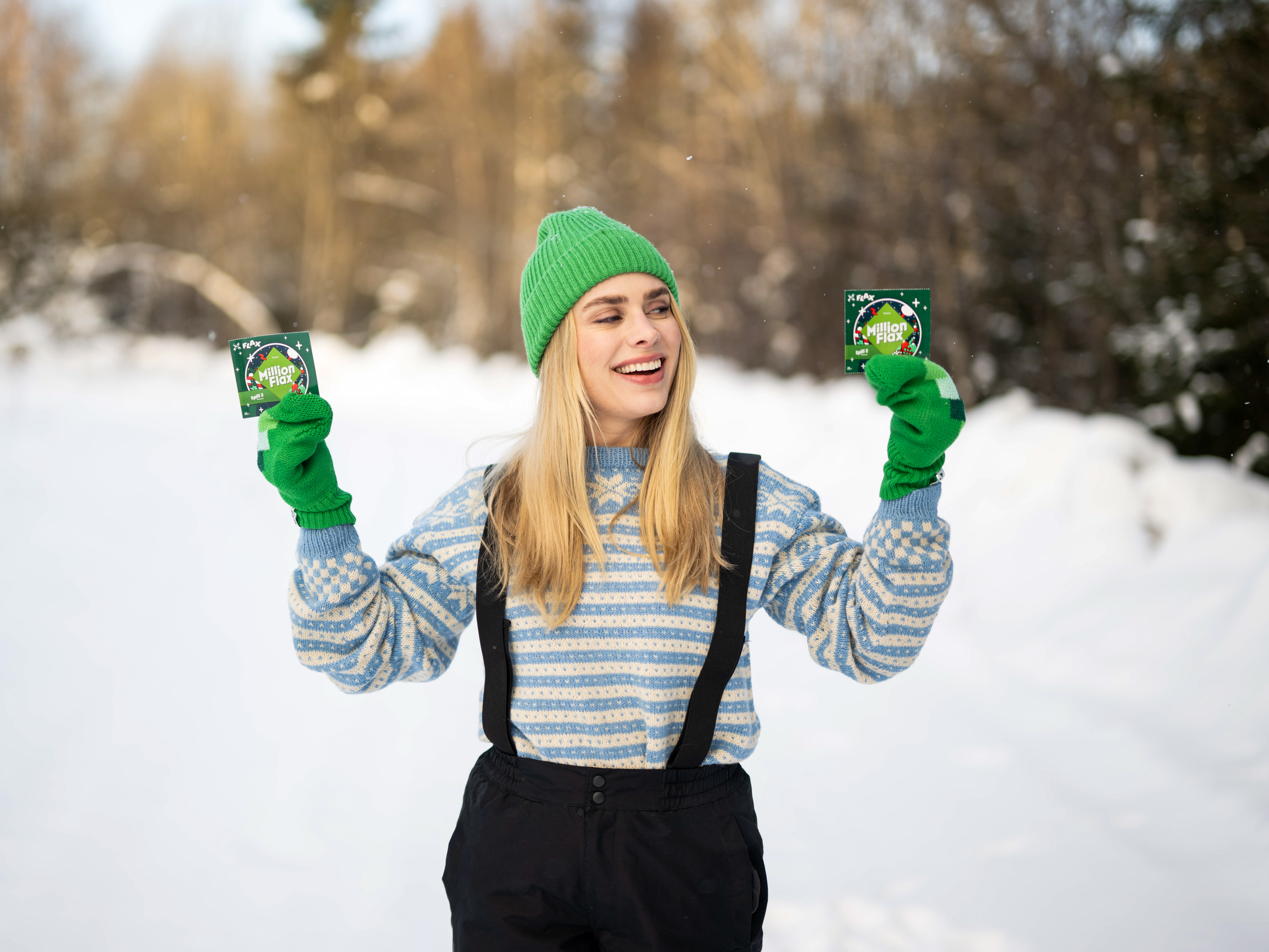 Blond kvinne i lyseblå strikkegenser, grønn lue og votter og sort overtrekksbukse med seler holder opp et MillionFlax i hver hånd, smiler og ser på det ene foran vinterskog og snø.