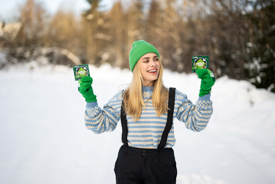 Blond kvinne i lyseblå strikkegenser, grønn lue og votter og sort overtrekksbukse med seler holder opp et MillionFlax i hver hånd, smiler og ser på det ene foran vinterskog og snø.