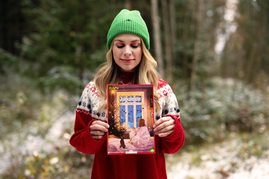 Blond kvinne med grønn lue og rød strikkegenser står foran vinterskog og holder opp Flax Julekalender foran brystet med begge hender mens hun kikker smilende ned.