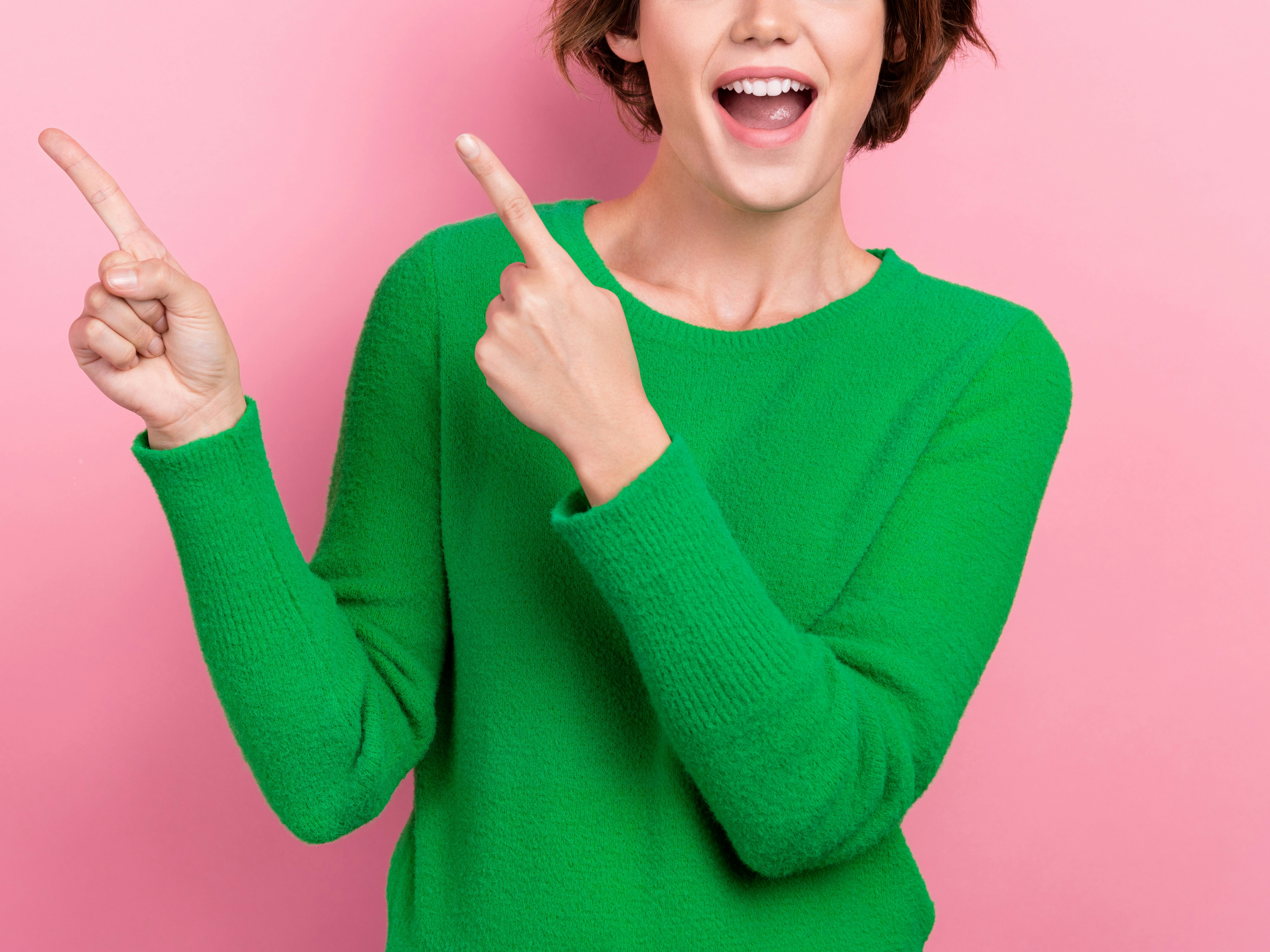 kvinne i grønn genser som smiler og peker med pekefingerne opp i luften. Rosa bakgrunn