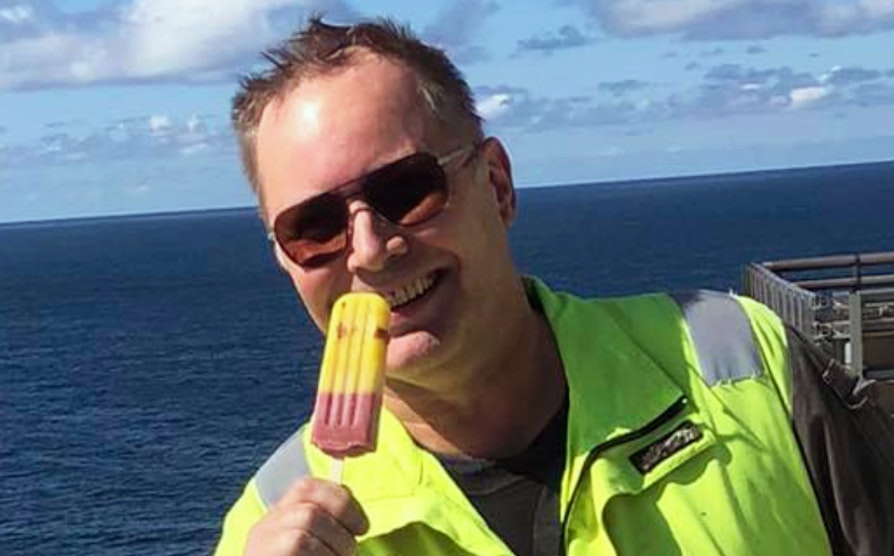 Portrett av Nabolaget-vinner Svein Arne Yven iført gul arbeidsvest og solbriller mens han smiler inn i kameraet og holder opp en is. I bakgrunnen skimtes havet.