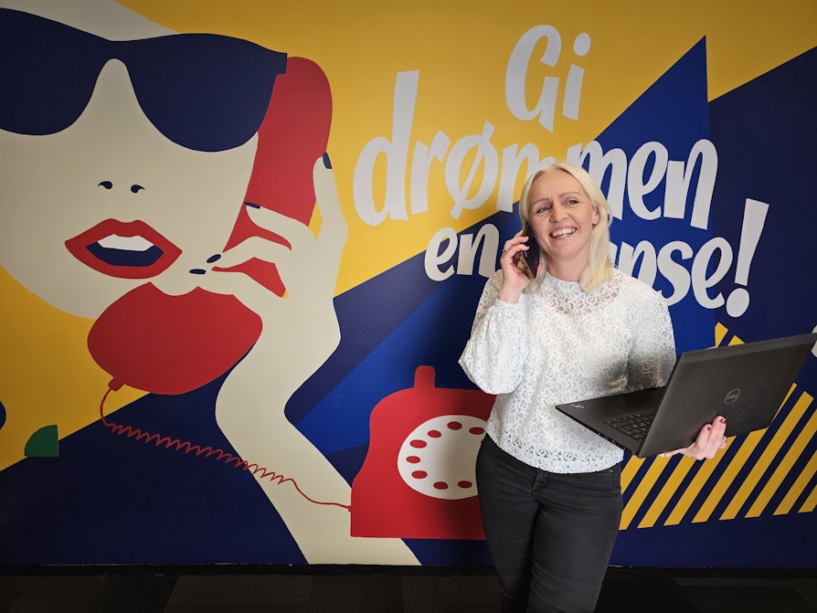 Bilde av Ingrid Roterud Mathisen i Norsk Tipping. Står foran en vegg der det står "Gi drømmen en sjanse". Snakker i telefonen og holder en bærbar pc.