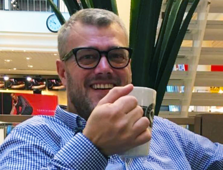 Portrett av Eurojackpot-vinner Erik Bakken i blå og hvit-rutete skjorte og sorte briller som smiler og holder opp en kaffekopp.