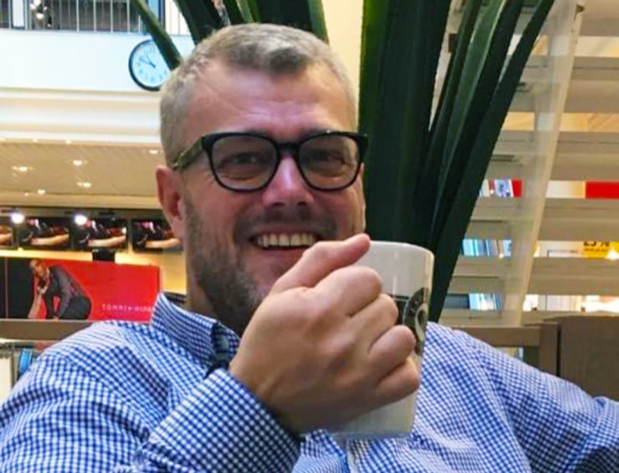 Portrett av Eurojackpot-vinner Erik Bakken i blå og hvit-rutete skjorte og sorte briller som smiler og holder opp en kaffekopp.
