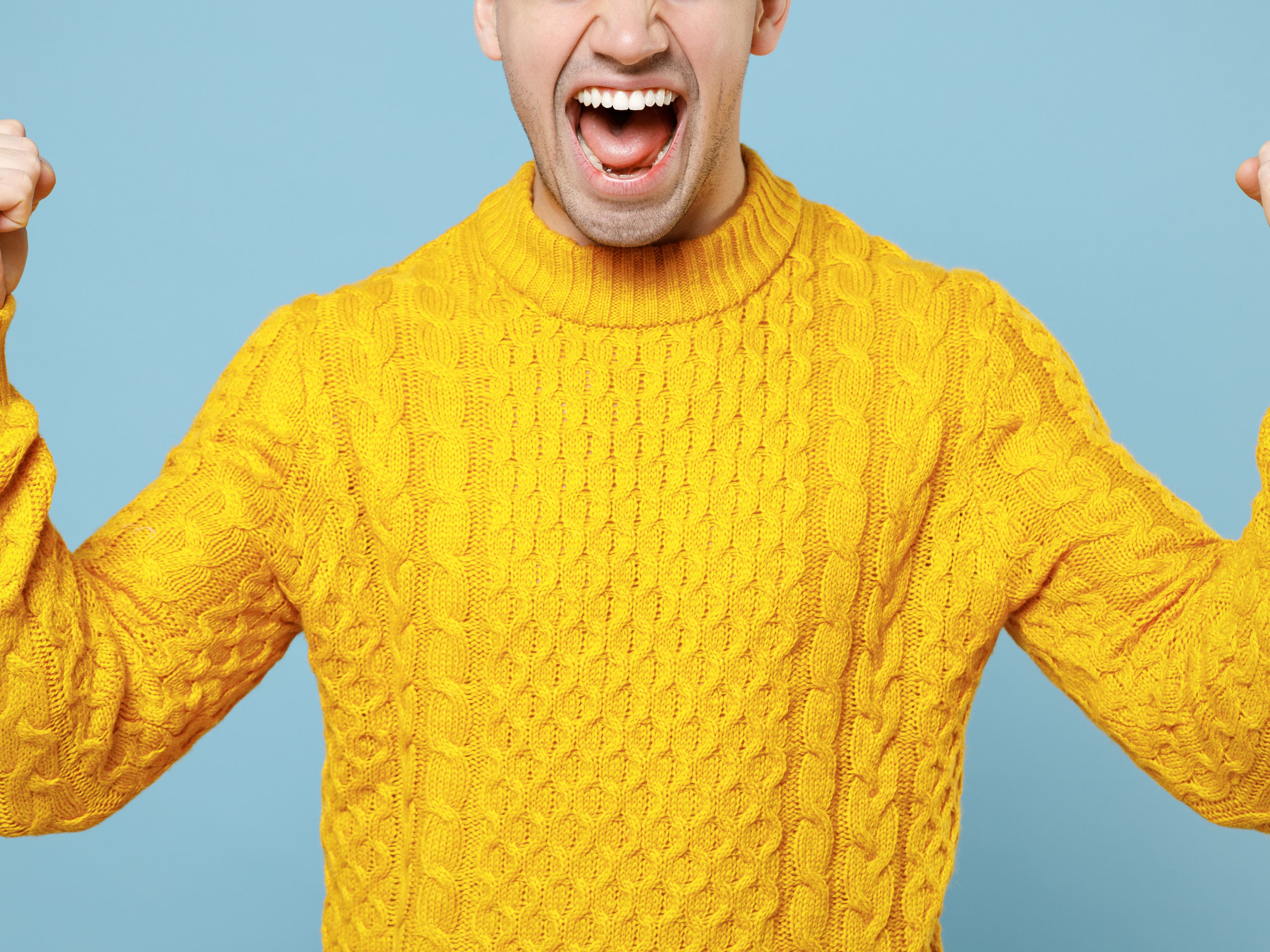 Utnsitt av overkropp og halve ansiktet til en mann som gaper jublende i gul strikkegenser og holder begge armene opp med hendene knyttet i seiersjubel, mot lyseblå bakgrunn.