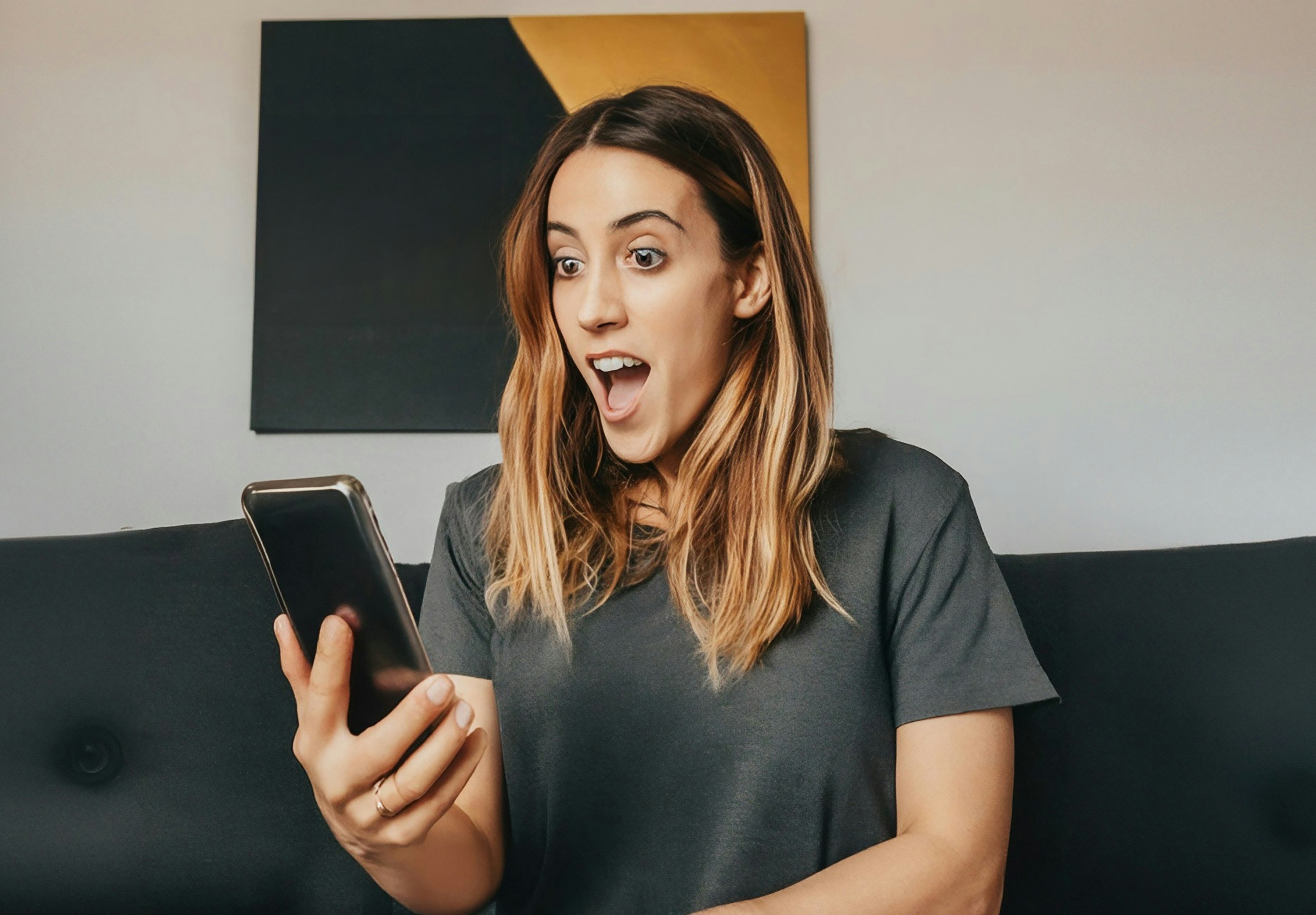 Bilde av en ung kvinne som ser overrasket på en mobiltelefon. Sitter i en sofa. Et bilde på veggen bak, der tallet 330 står. Skal illustrere at førstepremiepotten i Eurojackpot er på ca. 330 millioner kroner.