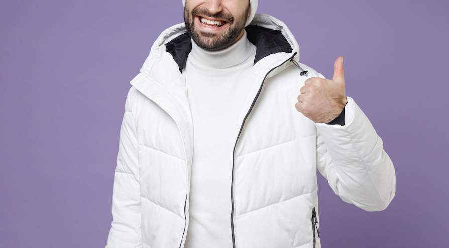 Utsnitt av halve ansiktet og overkroppen til mann med mørkt skjegg, hvit lue, pologenser og jakke, som smiler og viser tommel opp i kamera mot lilla bakgrunn.