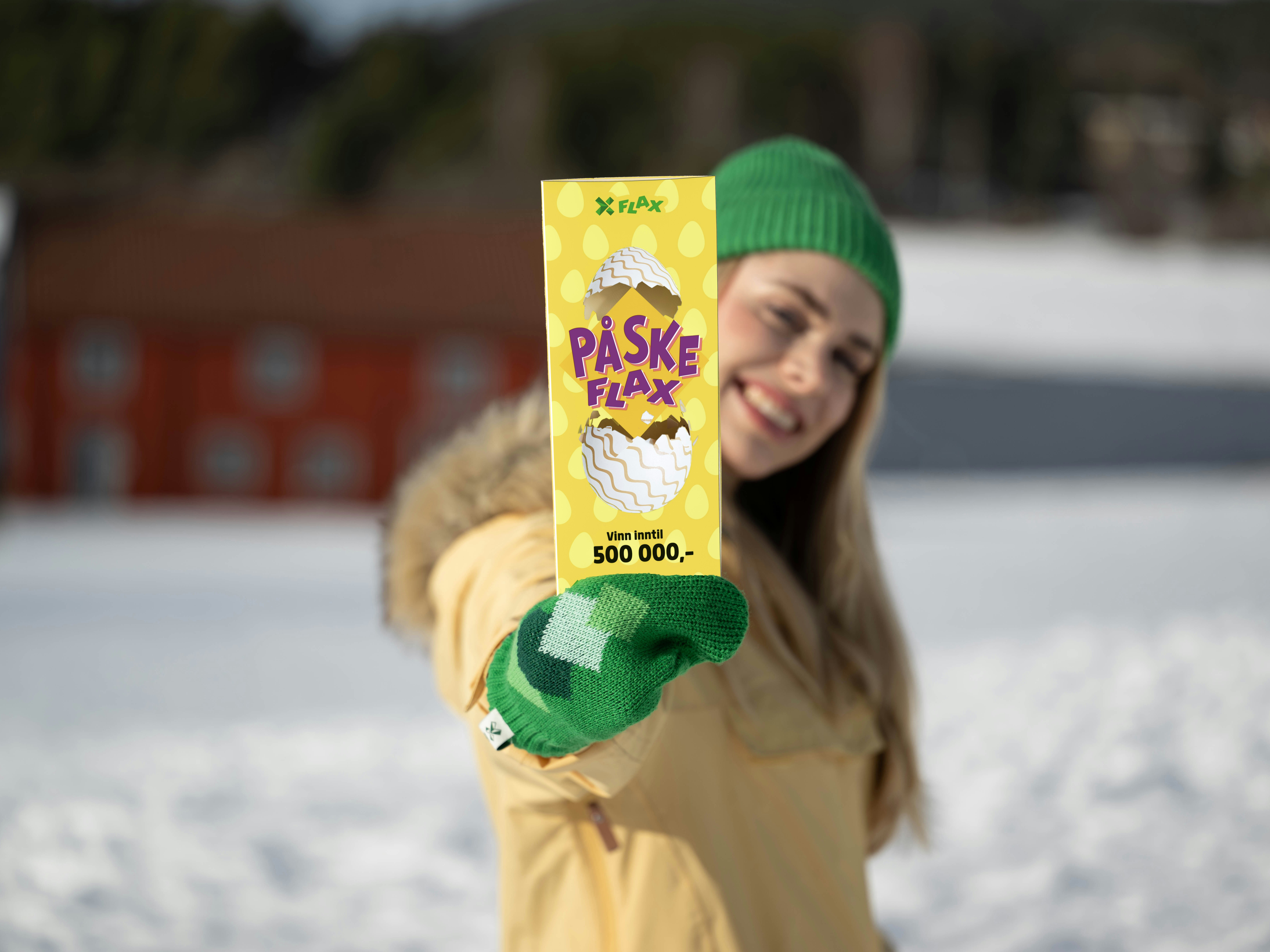 Kvinne med blondt langt hår, grønn lue og votter og gul anorakk holder frem et PåskeFlax og smiler  foran vinterlandskap.