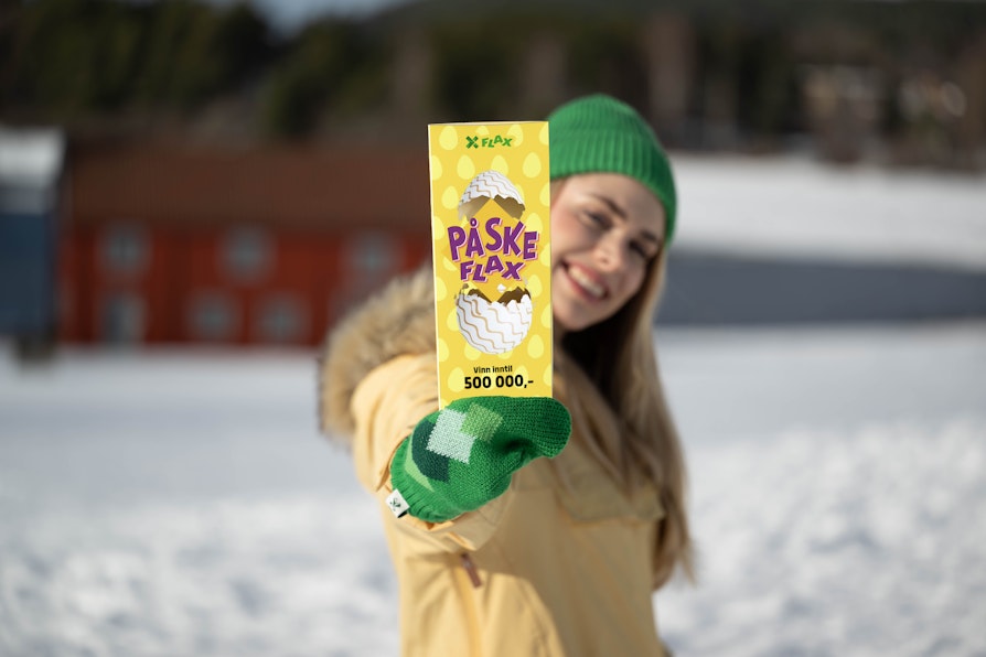 Kvinne med blondt langt hår, grønn lue og votter og gul anorakk holder frem et PåskeFlax og smiler  foran vinterlandskap.