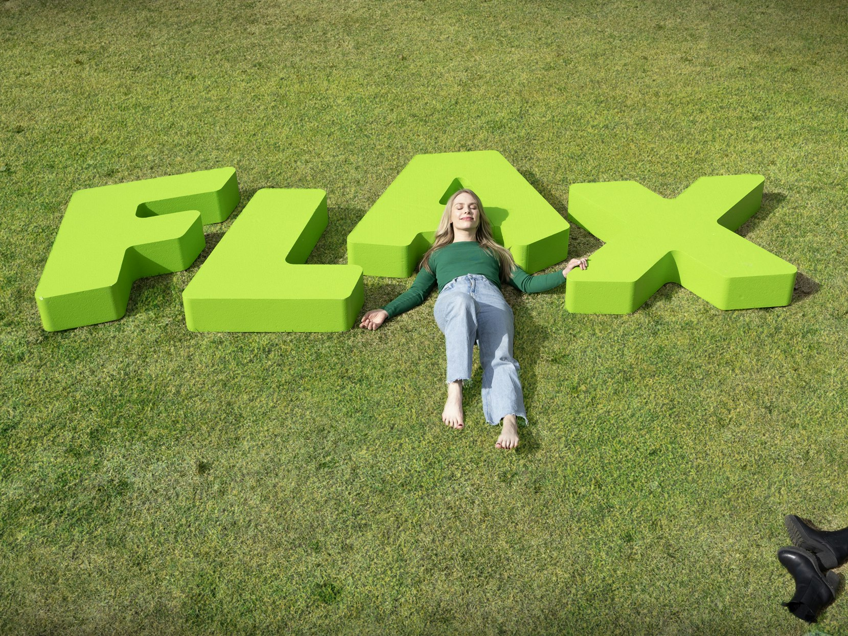 Kvinne ligger i gresset med og på bokstavene FLAX.
