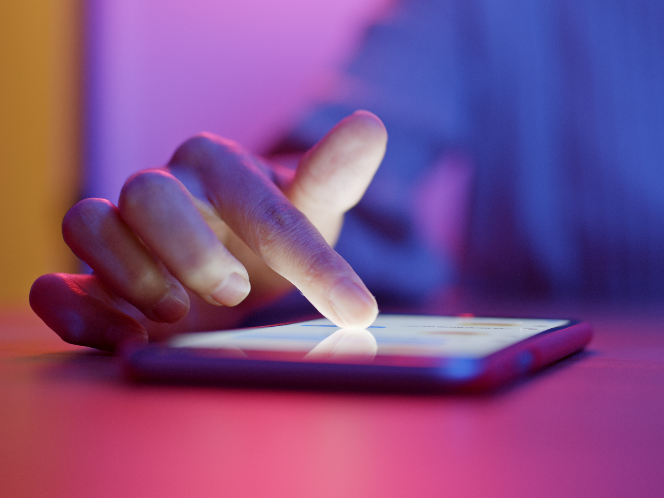 Nærbilde av mobiltelefon som ligger på et rosa bord med en hånd som trukker på den. Uskarp lilla, rosa og gul bakgrunn.