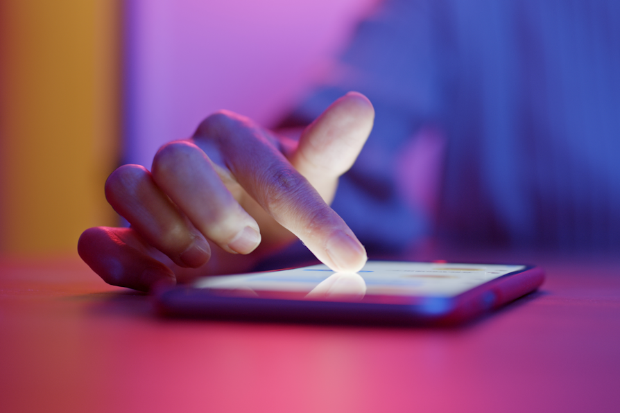 Nærbilde av mobiltelefon som ligger på et rosa bord med en hånd som trukker på den. Uskarp lilla, rosa og gul bakgrunn.