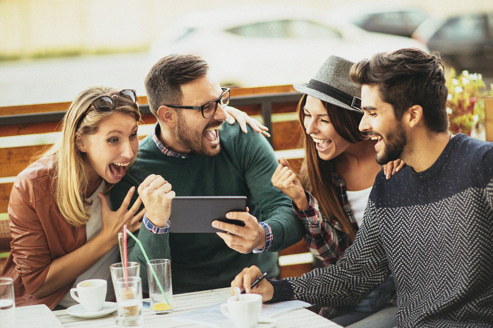 Bilde av fire unge mennesker. To kvinner og to menn. Sitter rundt et kafébord utendør og ser på en mobiltelefon. Er veldig glade over det de ser på telefonen.