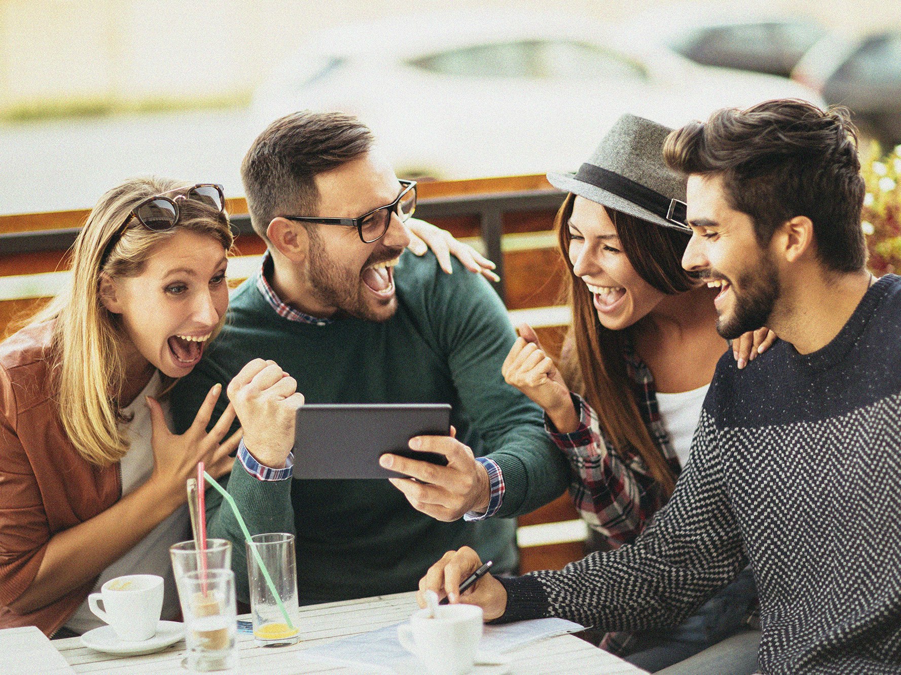 Bilde av fire unge mennesker. To kvinner og to menn. Sitter rundt et kafébord utendør og ser på en mobiltelefon. Er veldig glade over det de ser på telefonen.