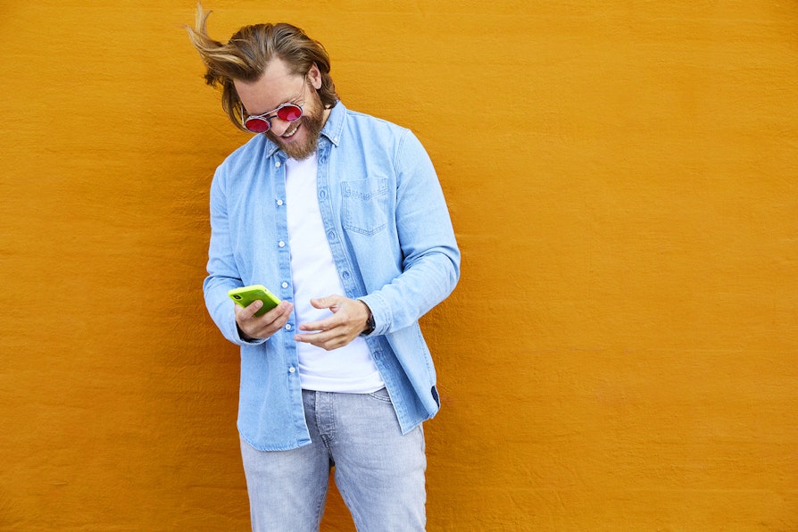 Bilde av en mann i 30-årene som ser på en telefon. Står foran en oransje vegg. Bildet brukt til Nabolaget-sak.