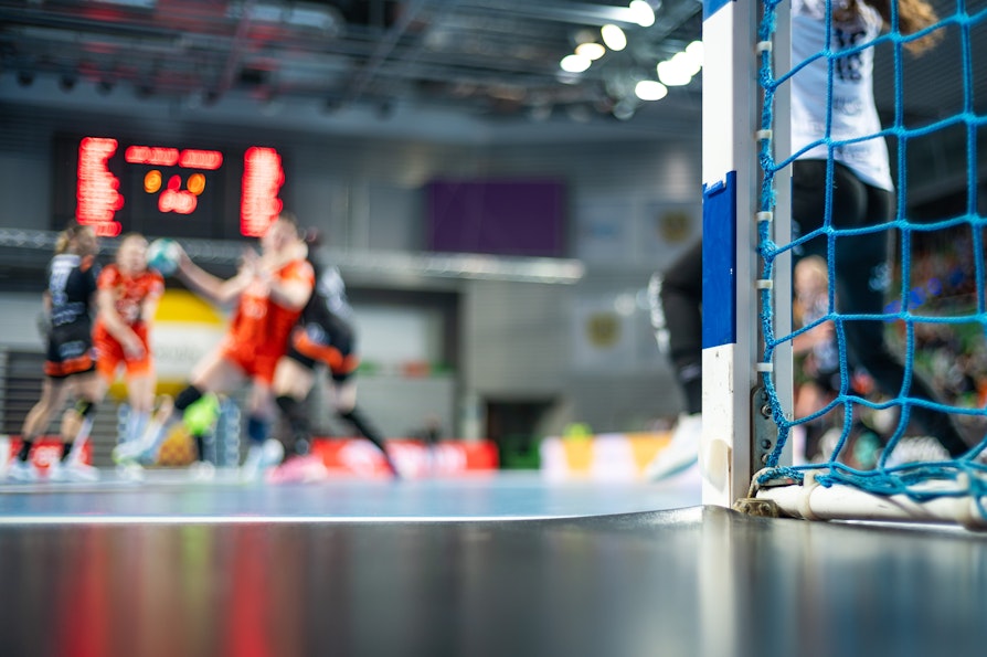 Bilde fra en håndballhall. Uskarpe spillere i bakgrunnen. Skarp målstolpe.