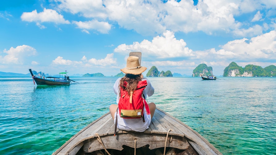 Bilde av kvinne på en båt ute på vannet i Thailand. Ryggen til. Rød ryggsekk. Noen båter rundt.
