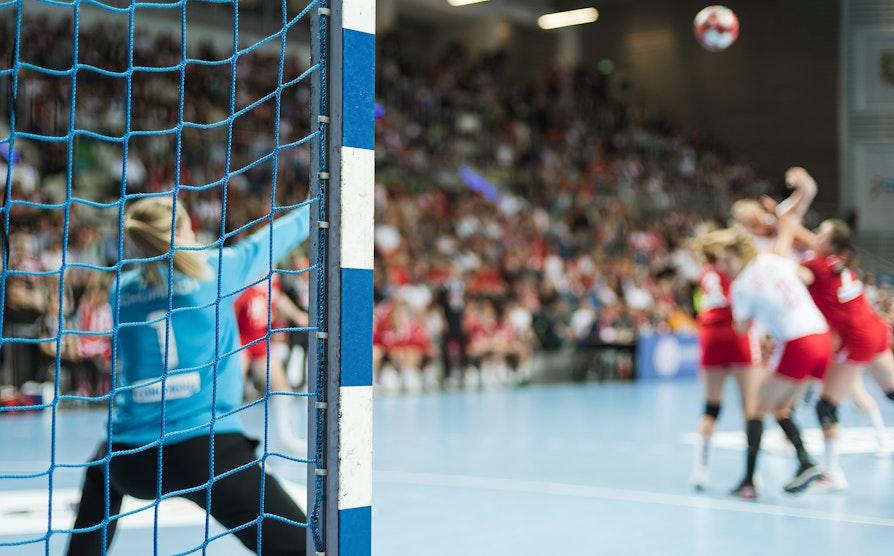 Bilde fra en håndballhall men håndballspillere. Uskarpe spillere i bakgrunnen. Skarp målstolpe.