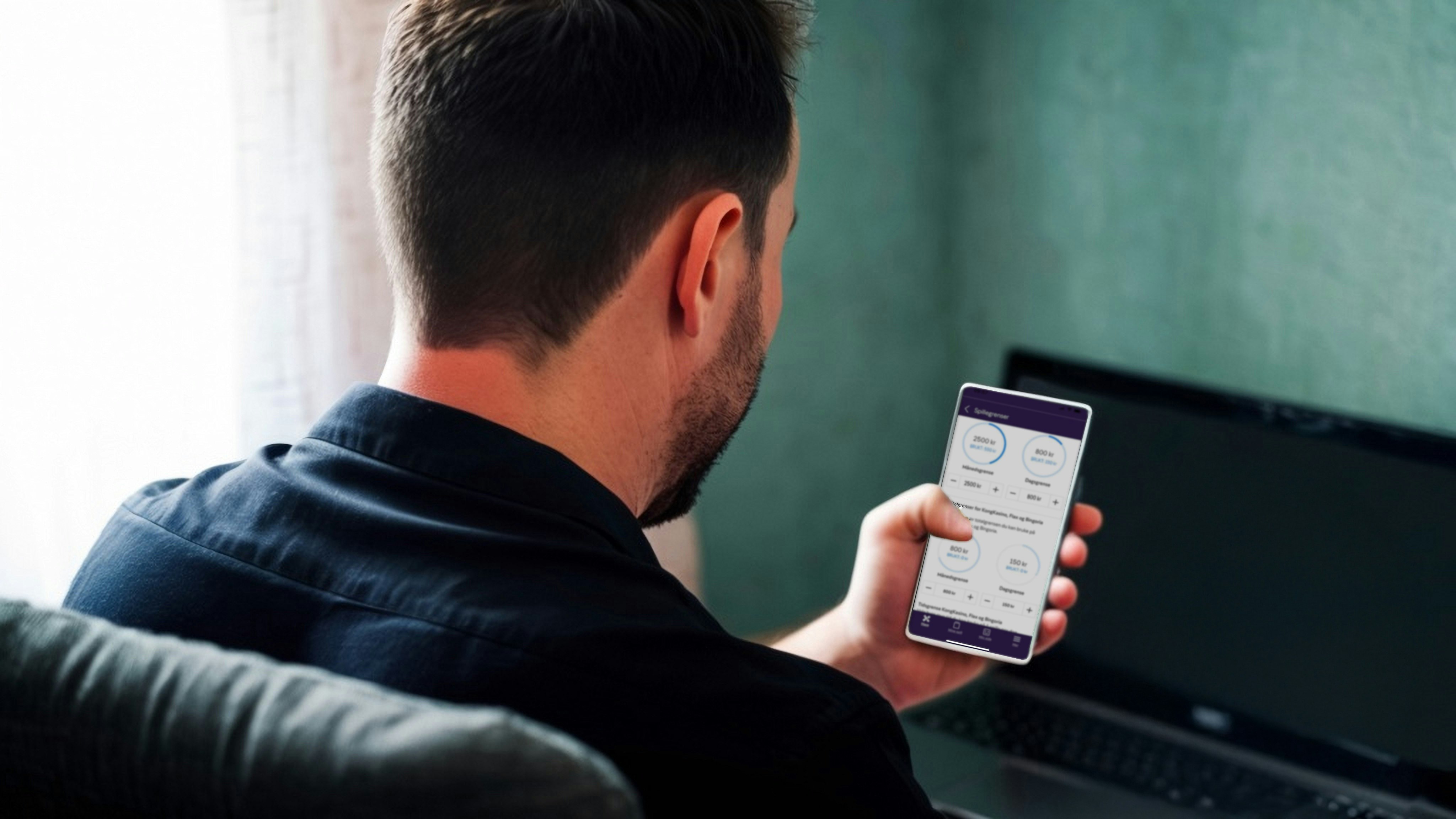 Bilde av mann, tatt bakfra. Ser på en telefon. PC i bakgrunnen.