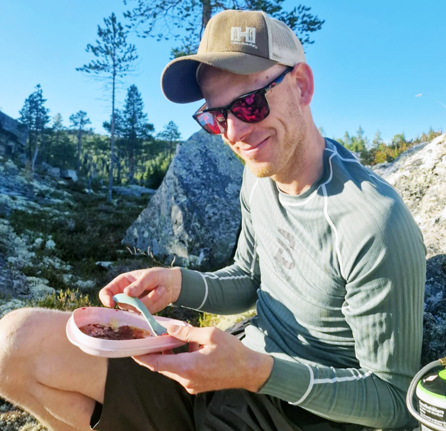 Extra-vinner Daniel Bakken med caps og solbriller, sitter ute i skogen og spiser mens han smiler.