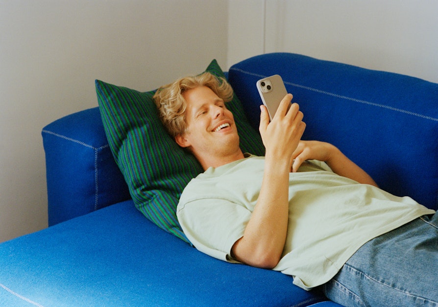 Blond mann ligger på ryggen i en blå sofa mens han smiler og ser på mobilen han holder i hånda.