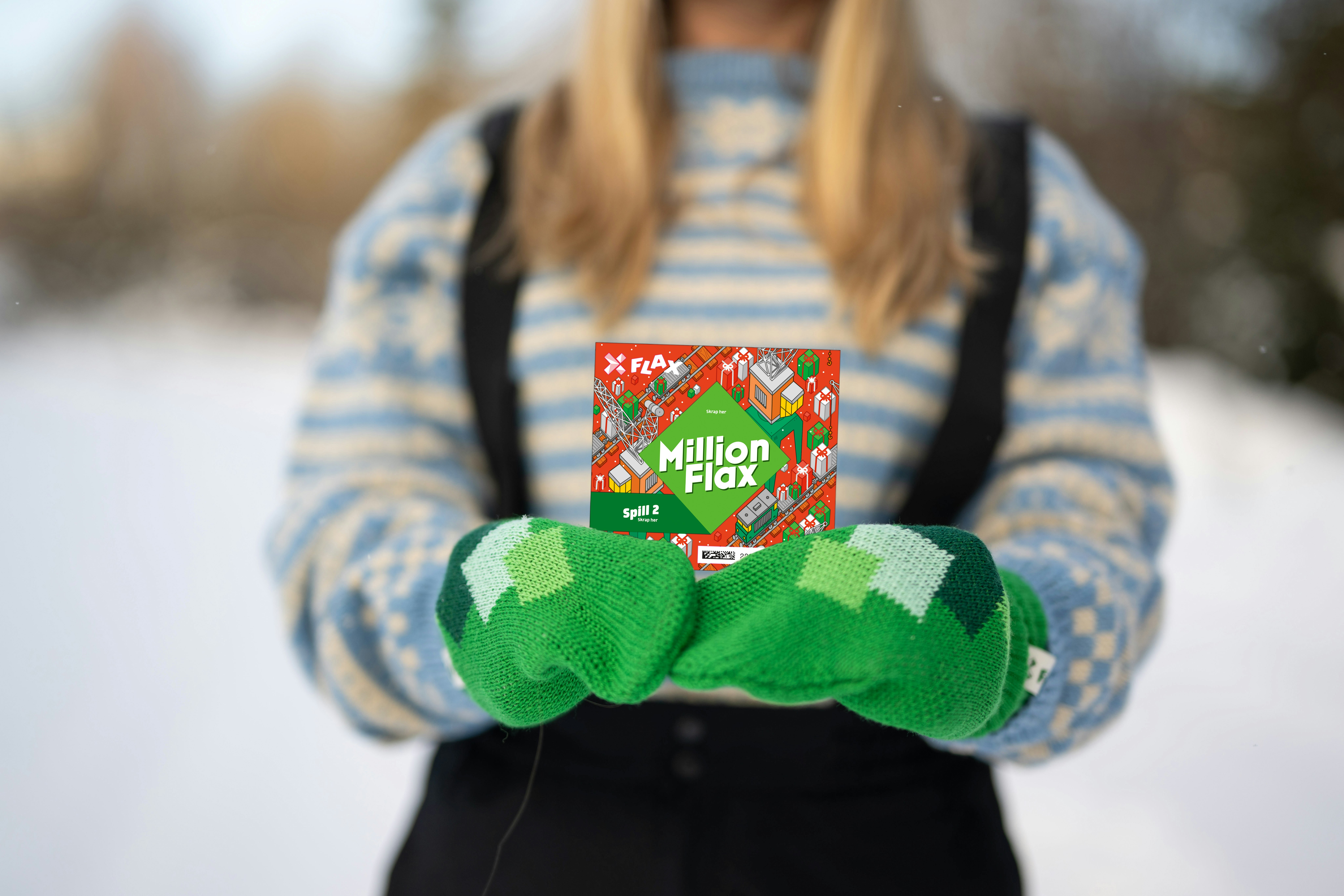 Utsnitt fra skulder til liv på kvinne med blondt hår, blåmønstret strikkegenser og grønne votter som holder et MillionFlax i begge hender foran seg.