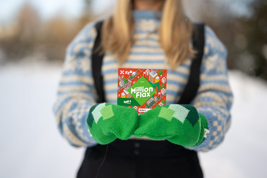 Utsnitt fra skulder til liv på kvinne med blondt hår, blåmønstret strikkegenser og grønne votter som holder et MillionFlax i begge hender foran seg.