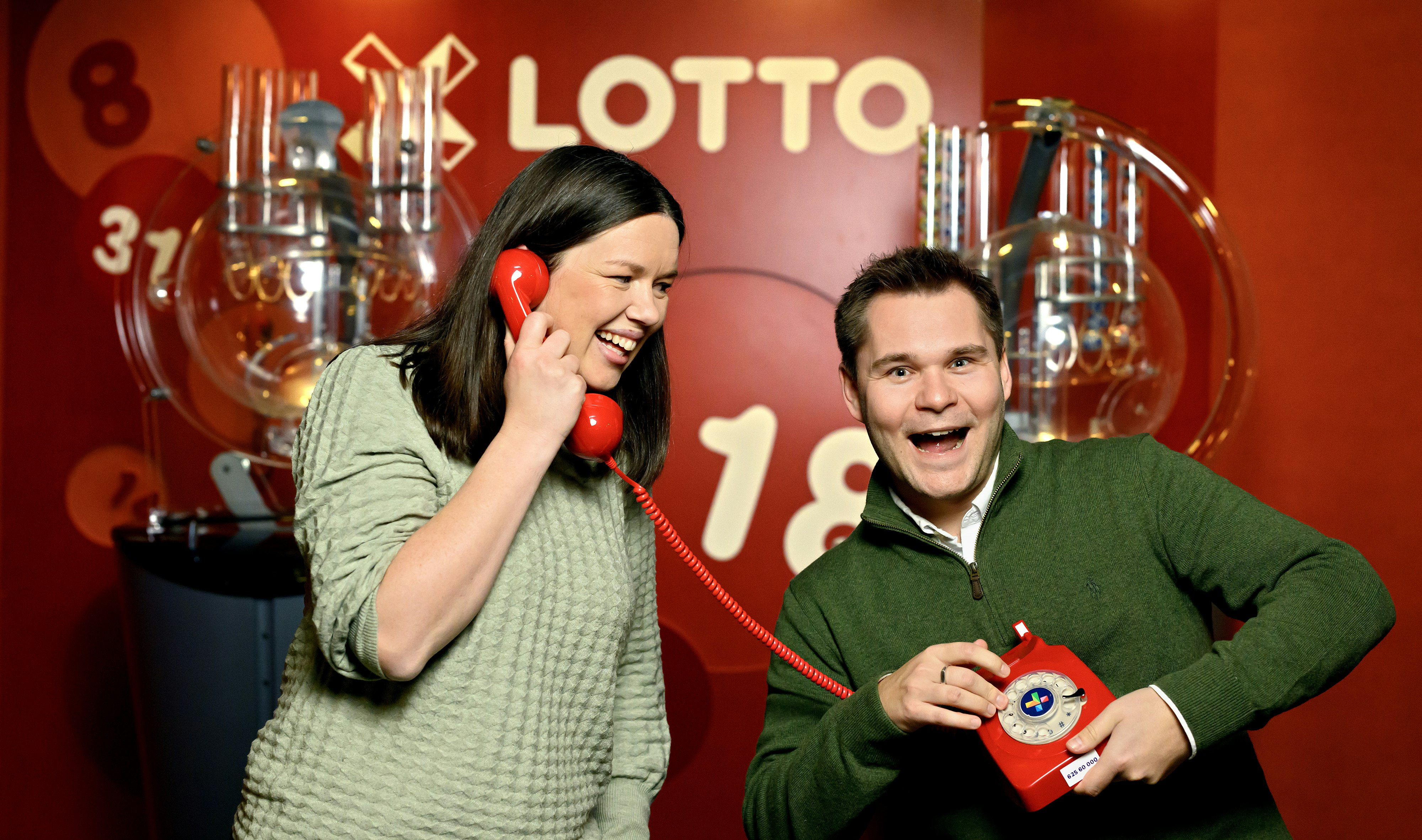 Bilde av Ingvill Kaasin Montgomery og Magnus Solberg Nygård i Norsk Tipping. Ringer vinnere i Supertrekning i Lotto 21. desember 2024. Står med en gammeldags telefon foran en rød Lotto-vegg. Det er også to trekningsmaskiner i Lotto i bakgrunnen.