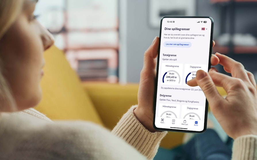 Blond kvinne i beige genser (ute av fokus og fra siden i skulderhøyde) sitter i gul sofa og holder olpp en mobil med begge hender som viser spillegrenser inne i Norsk Tipping-appen.