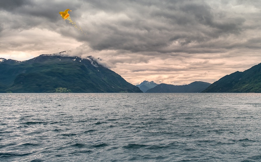 Fjord, fjell og gråskyet himmel i Stad Vestland, med jokerhatt på himmelen.