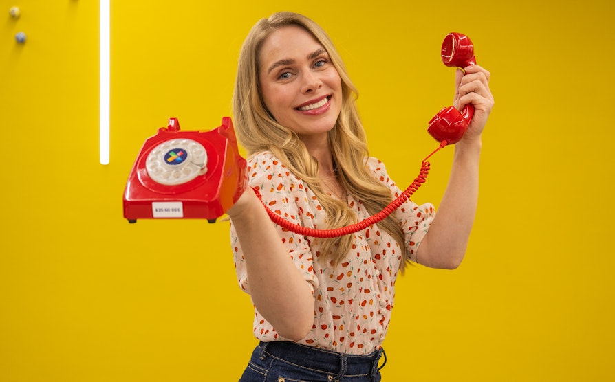 Blond kvinne holder opp en rød Lotto-telefon foran en gul vegg.